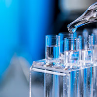 End of a pipette with a drop of clear liquid at the end over a rack of test tubes filled with clear liquid. 