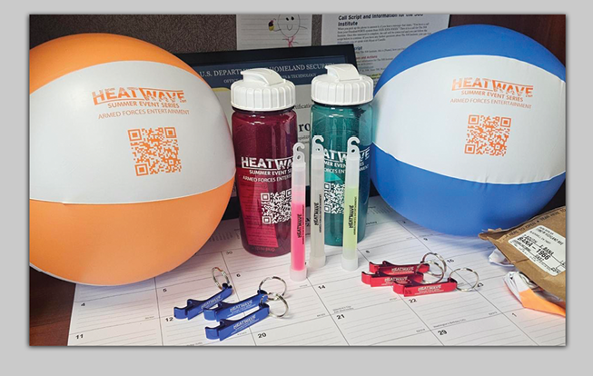 Merchandise for the HeatWave Summer Event Series sponsored by the Armed Forces Entertainment includes two beach balls, two waterbottles, three glow sticks (two yellow and one red), and six bottle opener keychains (three in blue and three in red) assembled on a table.